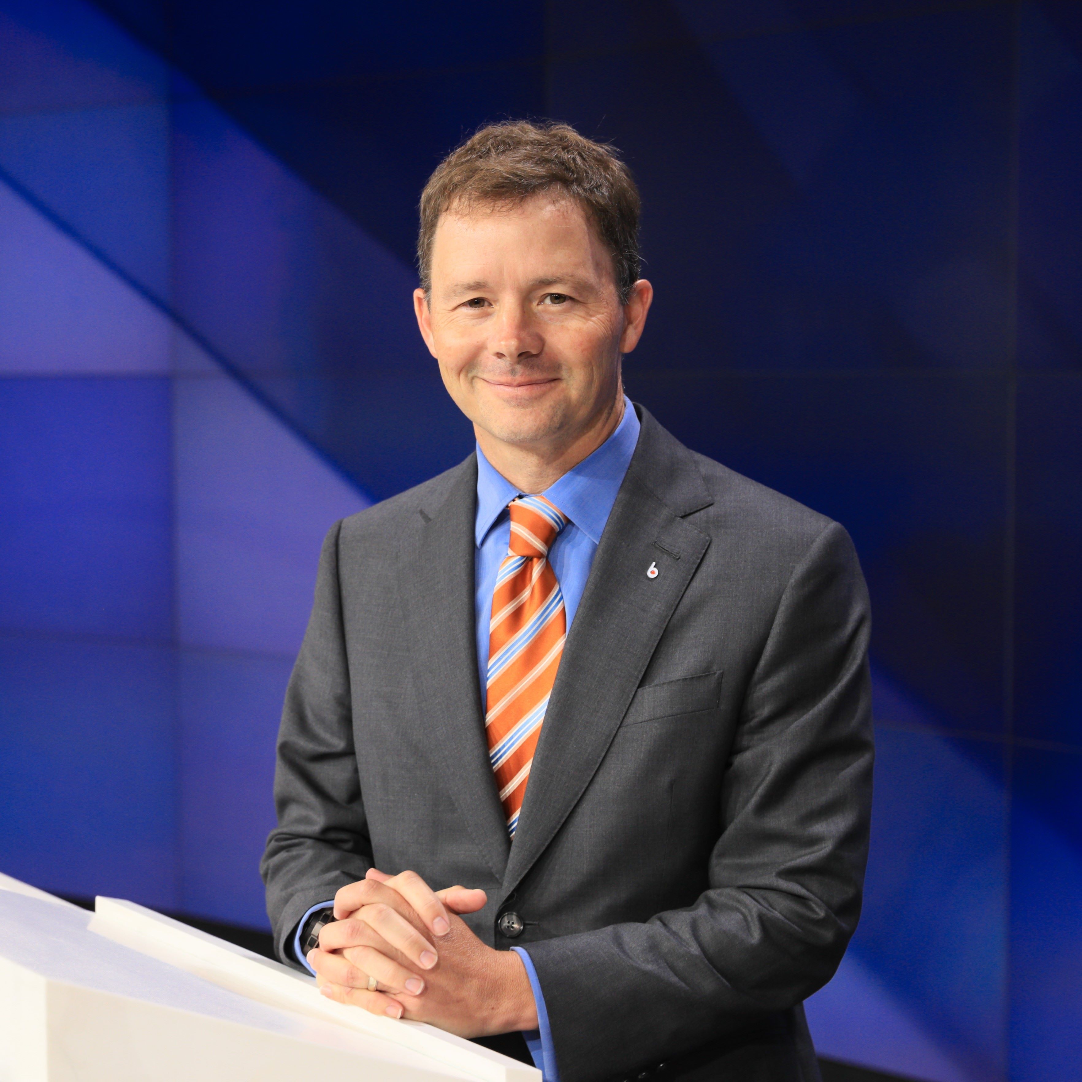 Jude Melville is in a dark suit at a white podium the background is blue.