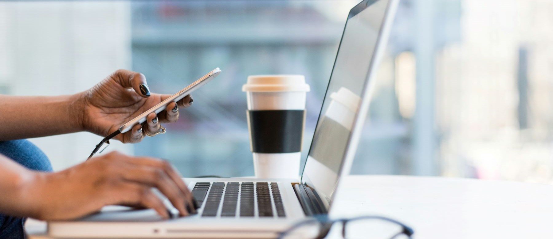 closeup of a person holding a smartphone in one hand and typing on a laptop with the other