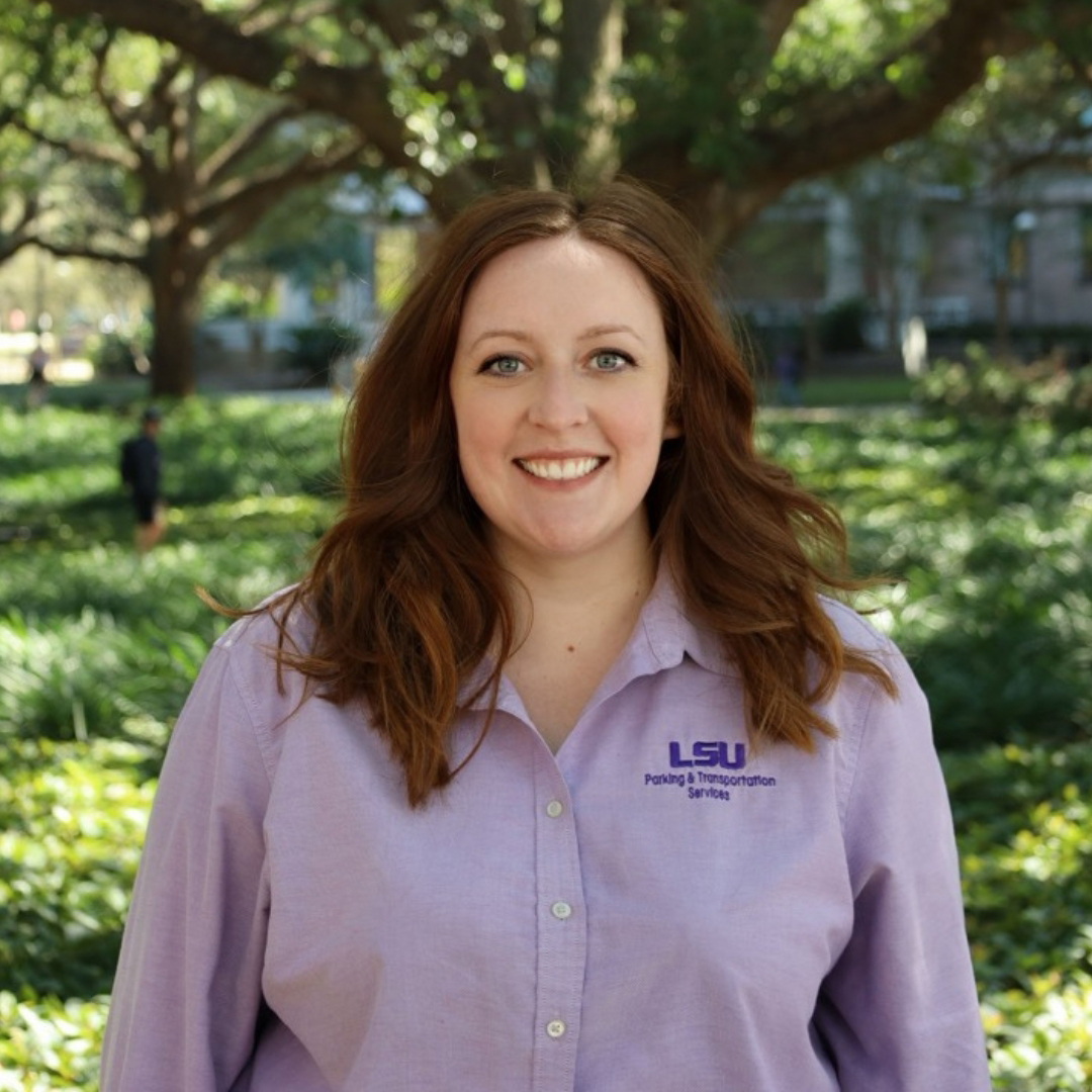 Woman in purple shirt smiling and standing in front of a tree and bushes